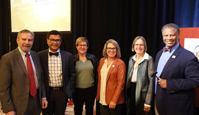 Group of cancer center leaders stands in front of a curtained background, Robert A. Winn holding a gavel